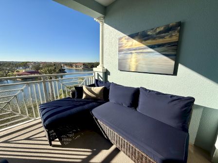A balcony with blue cushioned seating and a wall painting overlooks a scenic waterfront view under a clear blue sky.
