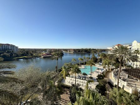 A scenic view featuring a waterfront resort with palm trees, a pool area, buildings, and a clear blue sky.