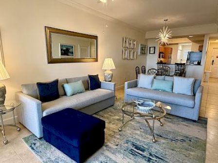 A modern living room with two sofas, a cushioned ottoman, glass coffee table, and decorative lamps, leading into a dining area and kitchen.