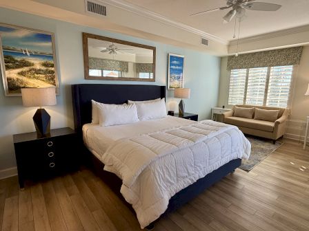 A cozy bedroom with a large bed, two side tables with lamps, wall art, a sofa, and a window with shutters letting in natural light.