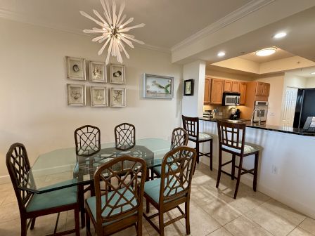 A dining area with a glass table and six chairs, artwork on the wall, and a kitchen in the background with a bar and stools.