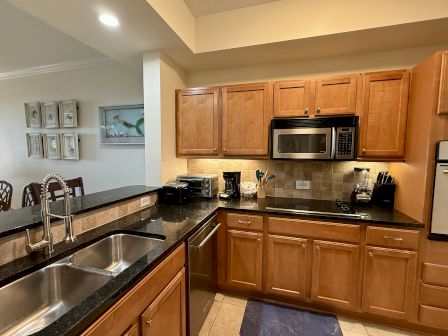 A kitchen with wooden cabinets, black countertops, a stainless steel sink, microwave, toaster, coffee maker, and decorative wall art.