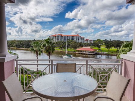 A scenic waterfront view from a balcony with a table and chairs, overlooking a river, palm trees, a yacht, and a large building.