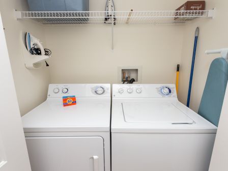 A laundry room with a washer, dryer, detergent, iron, ironing board, broom, and shelf.