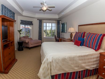 A cozy bedroom with a large bed, striped pillows, chaise lounge, TV, desk, and windows with a view, decorated in red and blue tones.