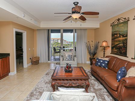 A cozy living room with a brown leather sofa, wall art, a ceiling fan, and a view of the balcony through sliding doors.
