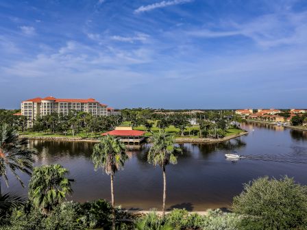A scenic view of a resort by a river with palm trees, a boat on the water, and clear blue skies.