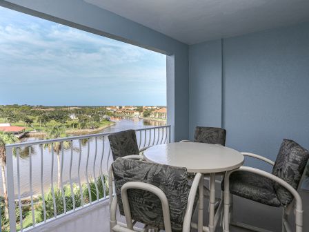A covered balcony with a round table and chairs overlooks a scenic view of a river, palm trees, and distant rooftops.