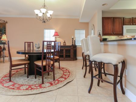 This image shows a dining room with a round table, chairs, a chandelier, a red rug, bar stools, and a kitchen area.