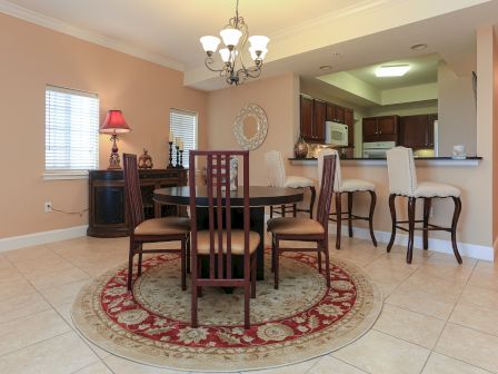A cozy dining area with a round table, four chairs on a patterned rug, a sideboard with lamp, and a breakfast bar with high stools.