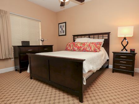 A cozy bedroom with a dark wood bed, floral pillow, nightstand with lamp, dresser, and framed art on the peach-colored walls.