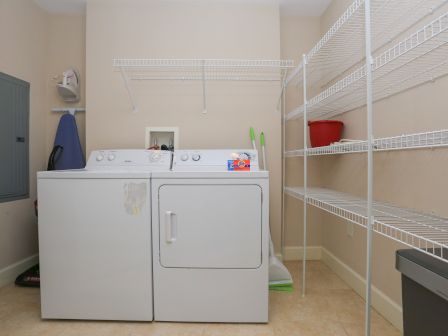 A laundry room with a washer, dryer, iron, ironing board, detergent, mop, broom, shelving, and a red bucket.