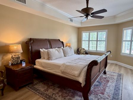 A cozy bedroom with a large wooden bed, side tables with lamps, a ceiling fan, and windows with shutters. A patterned rug covers the floor.