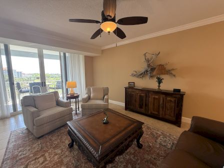 A living room with chairs, coffee table, ceiling fan, lamp, and wall decor. Large windows offer a view outside, creating a bright space.