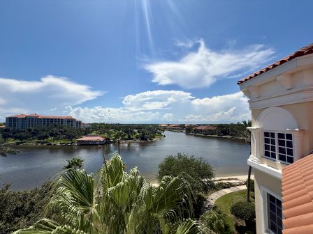 A scenic view featuring a waterway, palm trees, and buildings under a clear blue sky with white clouds in the background.