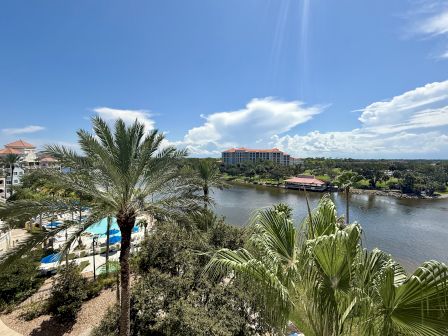 A scenic view of a river with palm trees, a distant building, and a clear blue sky with clouds.