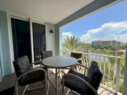 A balcony with a round table and four chairs overlooks a scenic view of palm trees and nearby buildings under a clear blue sky.