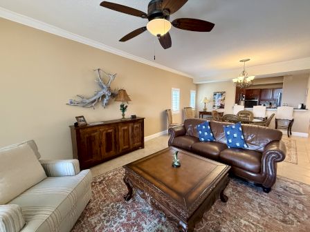 A cozy living room features a leather couch, armchair, wooden coffee table, and ceiling fan, with a glimpse of the kitchen area in the back.