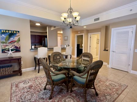 A cozy dining area with a glass table, wicker chairs, rug, chandelier, and a kitchen counter with stools in a neatly decorated room.