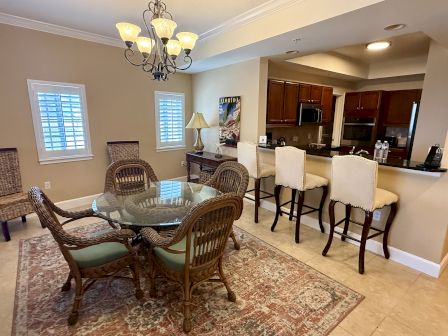 A dining area with a glass-top table, wicker chairs, kitchen bar with stools, chandelier, and artwork on the wall.