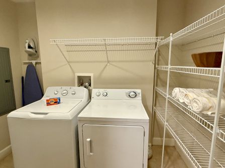 A laundry room with a washer, dryer, ironing board, wire shelves, towels, and a box on the washer.