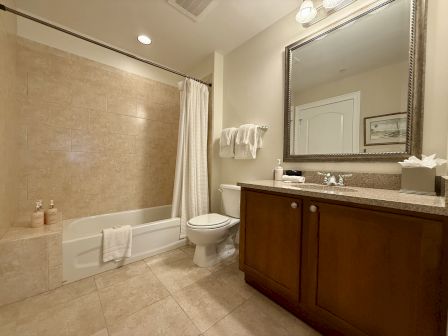 A bathroom with a bathtub, shower curtain, toilet, sink, mirror, and toiletries on a countertop, with beige tiles and wooden cabinets.