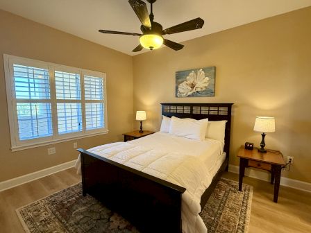 A cozy bedroom with a wooden bed, white bedding, nightstands, lamps, and wall art, illuminated by a ceiling fan light.