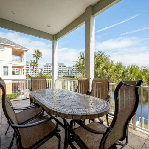 A patio with a large oval table and chairs overlooks a scenic view of water and palm trees under a clear blue sky.
