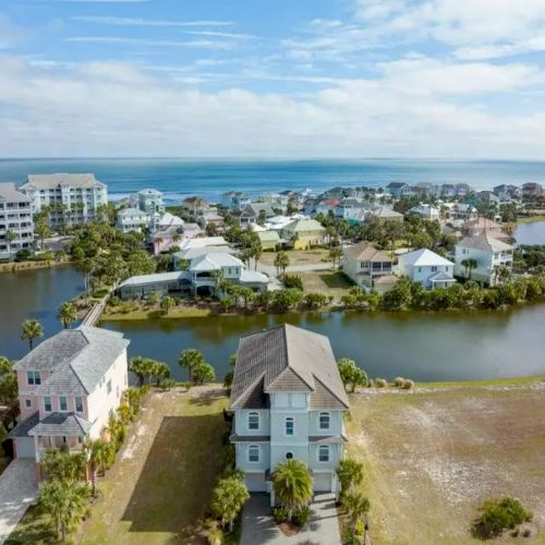 Aerial view of a coastal residential area with houses, waterways, and ocean in the background under a blue sky with clouds.