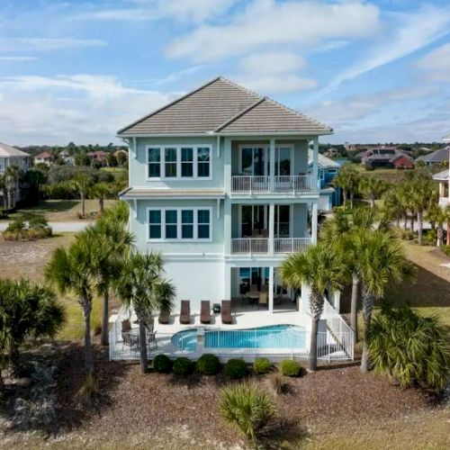 A multi-story house with a pool is surrounded by palm trees, and similar houses are visible in the neighborhood under a partly cloudy sky.