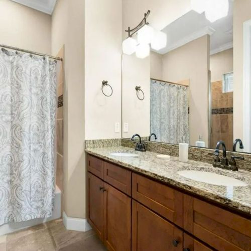 A bathroom with a granite countertop, double sinks, wooden cabinets, and a shower with a patterned curtain.