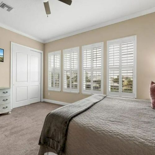 A bedroom with a bed, wall-mounted TV, dresser, and large windows with shutters. Neutral tones and ceiling fan complete the cozy space.