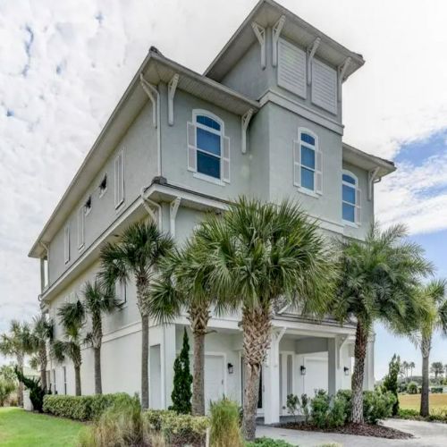 A tall, light-colored house with palm trees in front, situated in a grassy area under a partly cloudy sky.