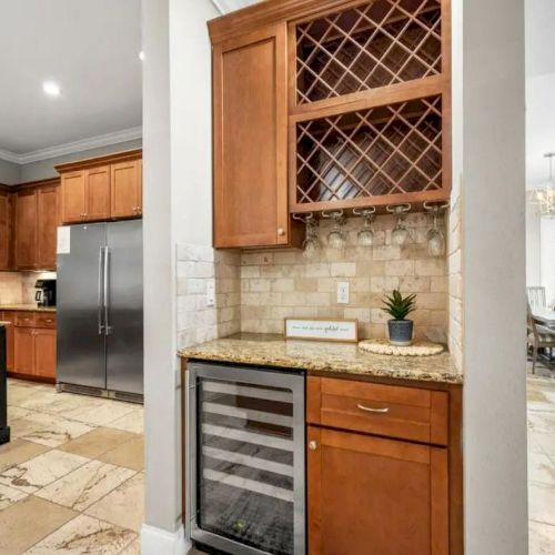 A kitchen area with wooden cabinets, wine storage, a mini fridge, and a dining area in the background.
