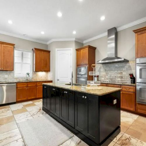 A modern kitchen with wooden cabinets, black island, stainless steel appliances, and tiled floor is shown in the image.
