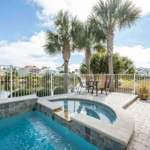 A sunny outdoor patio with a pool, spa, palm trees, and a dining set overlooks a scenic landscape with houses in the distance.