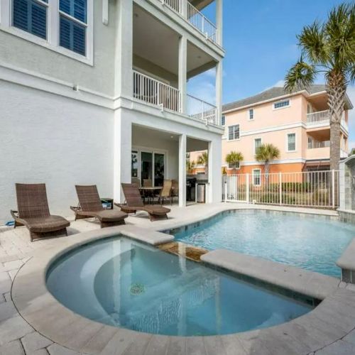 This image features a backyard with a pool and hot tub, surrounded by lounge chairs, palm trees, and homes in a sunny setting.