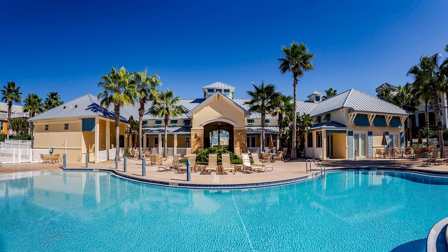 A resort-style pool with clear blue water, surrounded by palm trees and beige buildings under a bright blue sky.