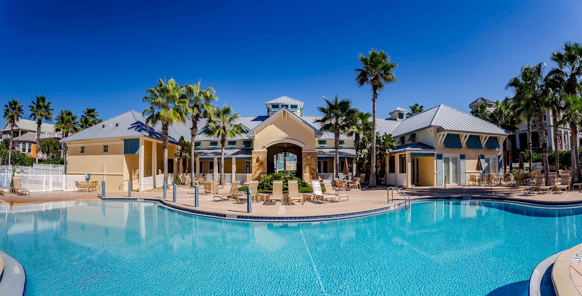 A resort-style pool with clear blue water, surrounded by palm trees and beige buildings under a bright blue sky.
