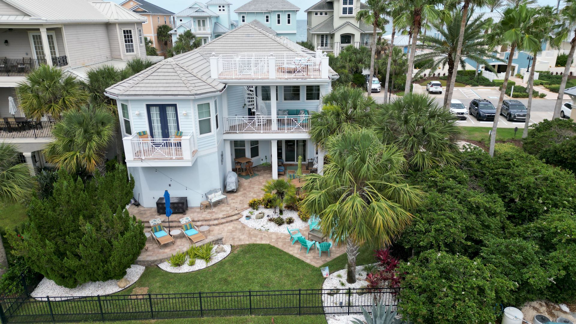 A coastal house with a multi-tiered balcony, surrounded by palm trees and a landscaped garden, with several parked cars nearby.