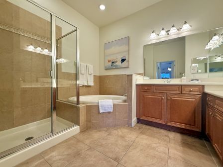 A bathroom featuring a glass shower, bathtub, brown cabinets, double sinks, and wall art, illuminated by ceiling and mirror lights.