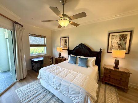 A cozy bedroom with a large bed, blue pillows, a ceiling fan, lamps, artwork, wooden furniture, a window, and sliding glass doors.