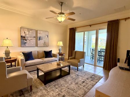 A cozy living room with a beige couch, armchairs, abstract art, ceiling fan, sliding glass doors, and a patio dining set visible outside.