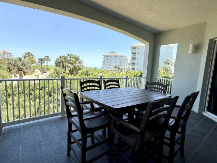 A covered balcony with a large wooden dining table and chairs overlooks lush greenery and buildings in the distance under a clear blue sky.
