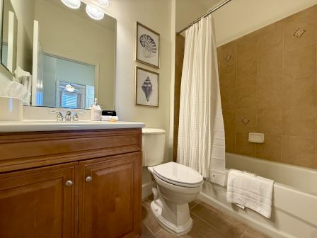 A bathroom features a sink, toilet, mirror, white shower curtain, and framed shell artwork on the wall above brown cabinetry.