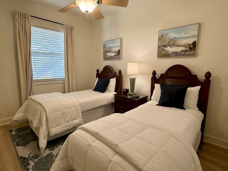 A cozy bedroom with two twin beds, white bedding, blue pillows, and paintings above. There's a lamp and clock on the nightstand.