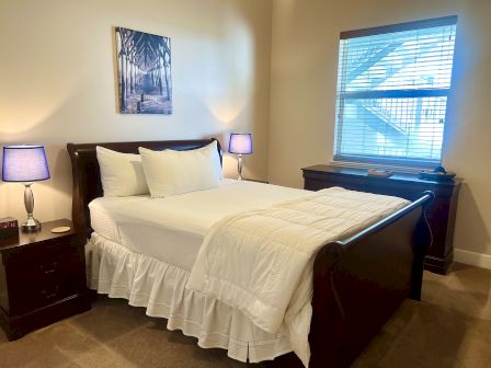 A neatly made bed with white linens, wooden furniture, blue lamps, a window with blinds, and a wall art piece above the bed.