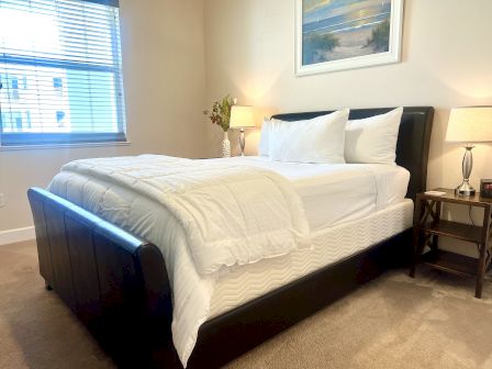 A tidy bedroom with a neatly made bed, two lamps on side tables, a window with blinds, and a framed landscape painting on the wall.