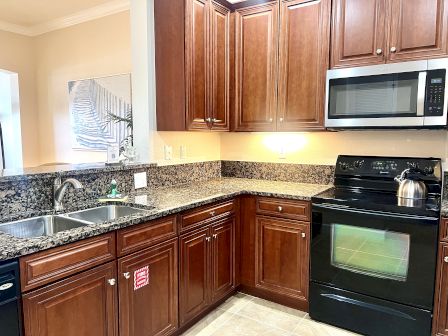 A kitchen with wooden cabinets, granite countertops, a double sink, microwave, electric stove, and a kettle.