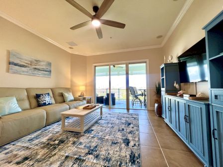 A cozy living room with a large sofa, patterned rug, and blue cabinets faces a patio with outdoor seating and scenic views.